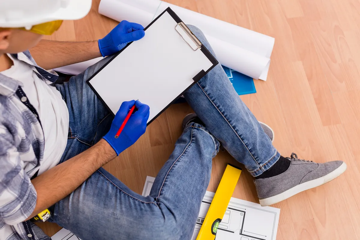 Taking notes on a clipboard while sitting on a wood floor with floor plans and a level